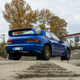 rear shot of a blue subaru coupe parked on a gravel lot. the photo is taken from an extremely low vantage point, showing off the rally-racing rear spoiler on the subaru's trunk lid, as well as the gold wheels and all-terrain tires